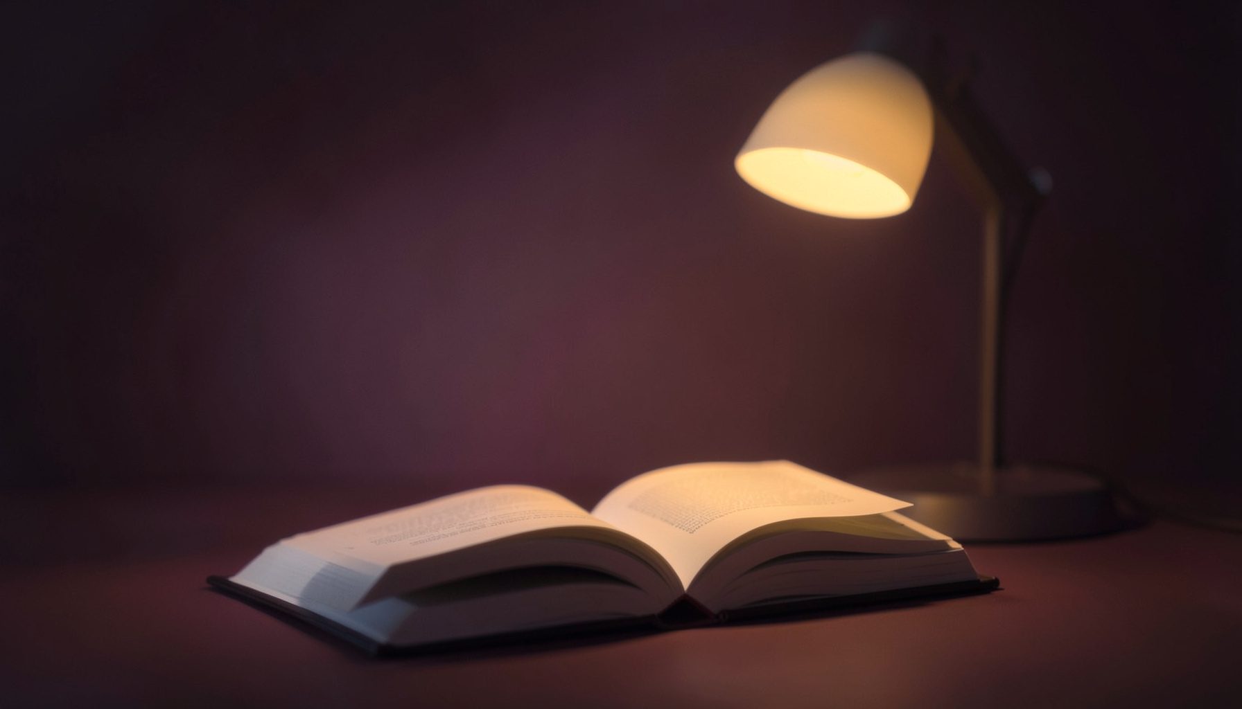 Loş bir ortamda açık duran bir kitap, yanında sıcak tonlarda bir masa lambası; koyu lacivert ve kehribar rengi atmosfer
